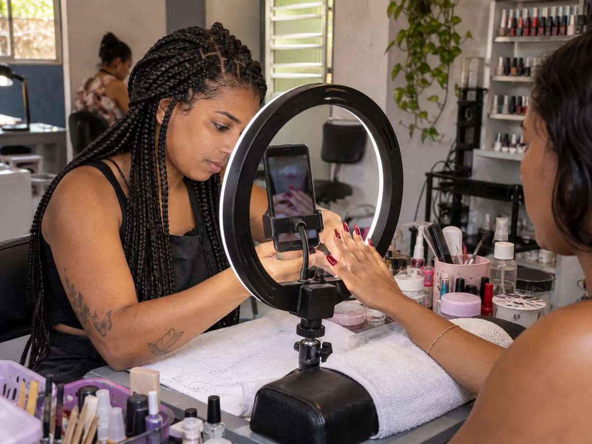 Manicure filma unhas recém-feitas com o celular preso no meio de uma luz circular